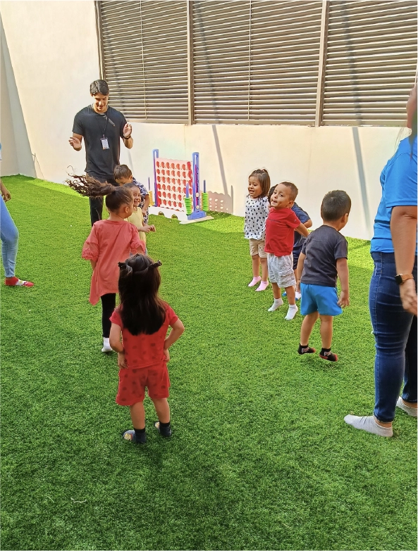 Un grupo de niños pequeños juega en el patio con pasto sintético bajo la supervisión del personal de CAYAM.